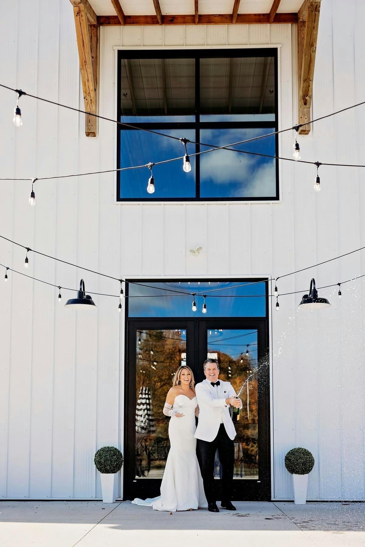 Picture of Bride and groom popping a bottle of shampain in front of building with string lights