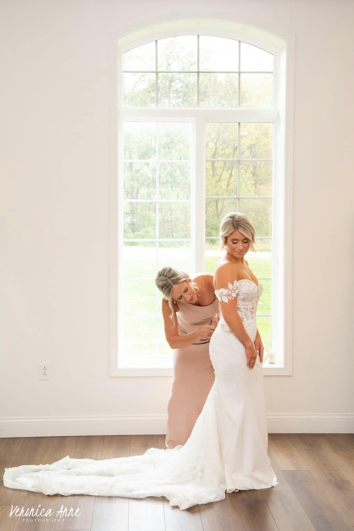 Picture of bridesmaid helping bride with her dress in front of big beautiful window