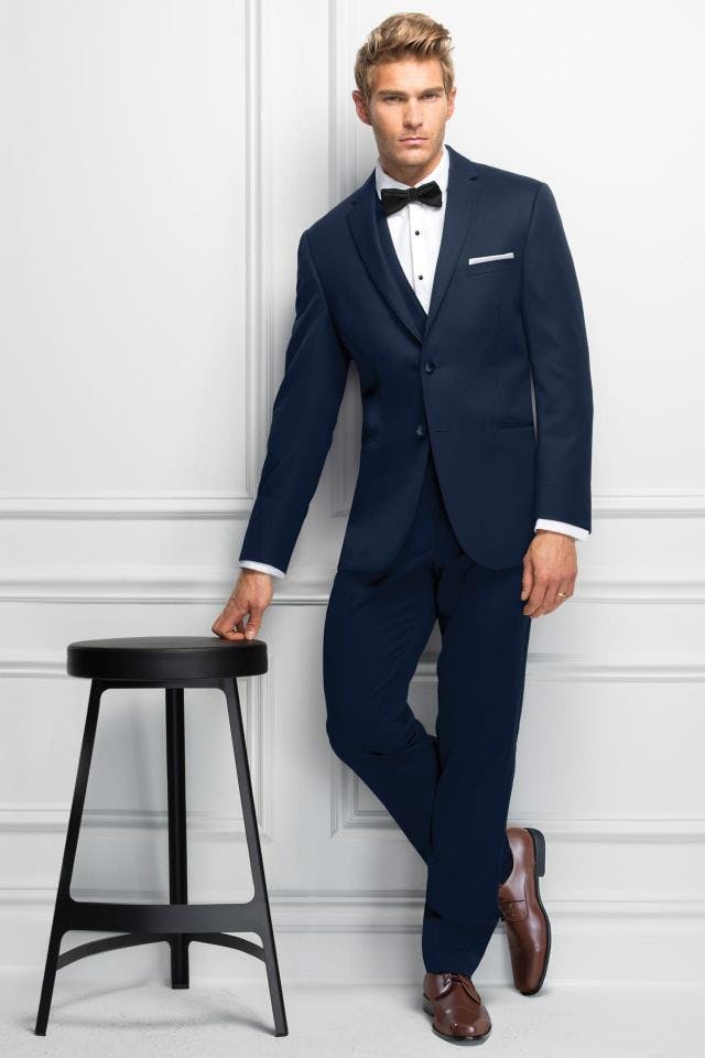 Picture of man in blue tux in front of white wall leaning on a stool